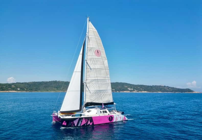 A white catamaran with a magenta hull under full sail on a blue gulf, a green headland behind