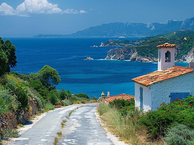 A narrow road down to a small white house above the sea, a rocky wooded cape beyond